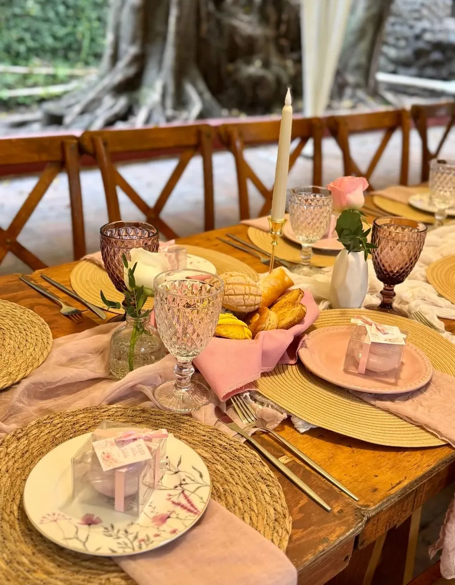 Mesa en tonos blush con canastas de pan dulce, velas y vajilla floreada bajo un árbol del jardín