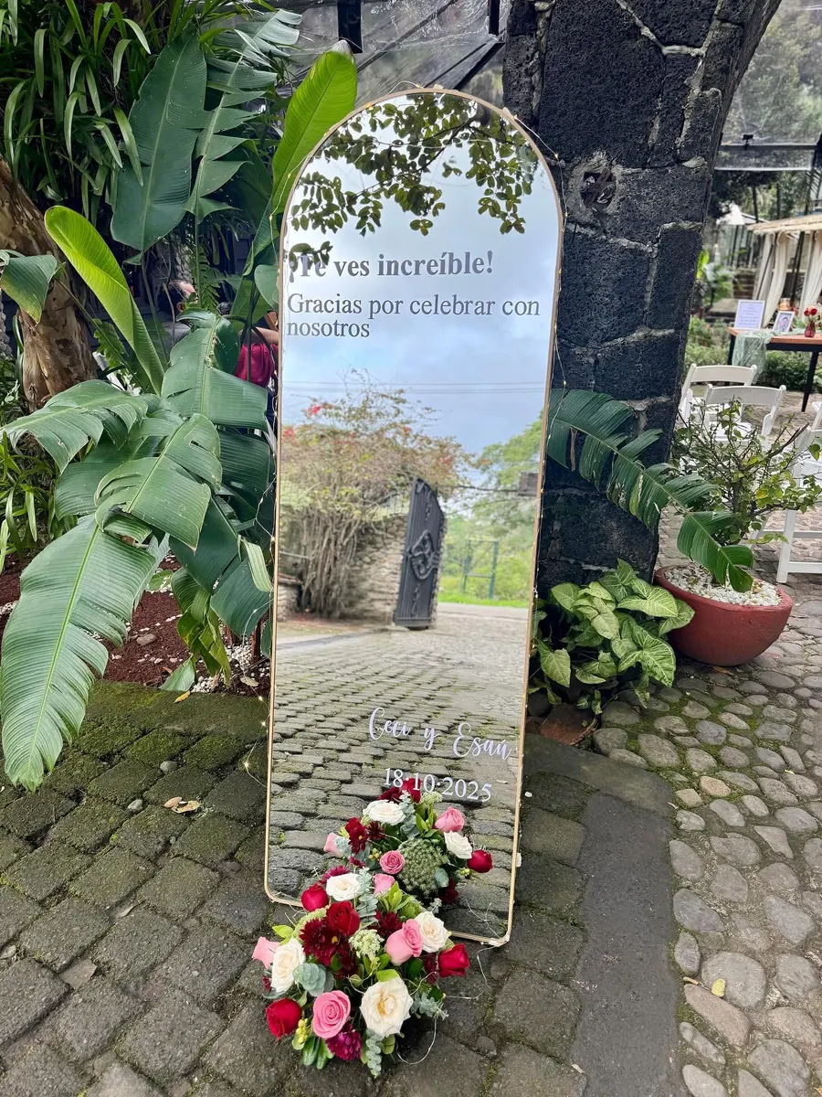 Espejo arqueado con el mensaje '¡Te ves increíble! Gracias por celebrar con nosotros' y flores al pie, en la entrada empedrada del jardín